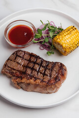 Overhead view of a delicious grilled striploin steak with grilled corn on the cob, pickled red onions, and a small bowl of barbecue sauce, placed on tabletop. Restaurant or cafe menu, cookbook recipe