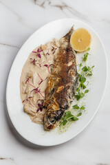 Delicious grilled fish served on a white plate with creamy sauce, lemon slice, microgreens, and red sprouts, creating an appetizing culinary presentation. Restaurant or cafe menu, cookbook recipe