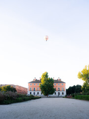 Morning in Madrid, Spain. A hot air balloon flies over the top of an old orange and white building in the city, on the edge of a park. The rising sun is casting an orange glow on the view.