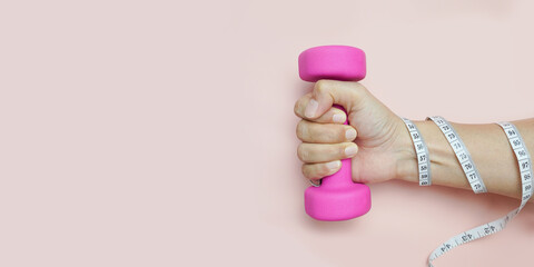 Woman's hand holds the dumbbell and measuring tape on a pink background with copy space. Creative concept for a healthy lifestyle.