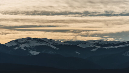 Cold snowed hills with pine trees forests over cloudy sky at sunset light in winter near the city 