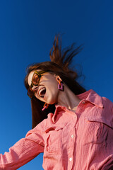 Excited woman expressing joy outdoors in a vibrant pink shirt with large earrings against a clear...