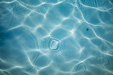 Close-up of transparent blue water with ripples, sun glare, and wave patterns on the surface, creating reflections and textures at the bottom. Outdoor abstract background.  