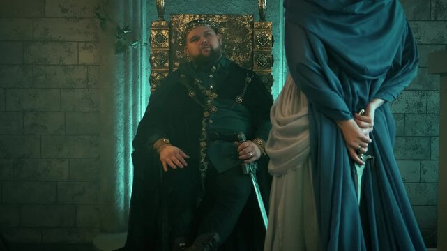 Fantasy Evil tyrannical man king sits medieval royal throne. Woman Queen, blue dress cape, holding in hands steel knife dagger. Women revenge. Girl dangerous princess back rear view old style castle