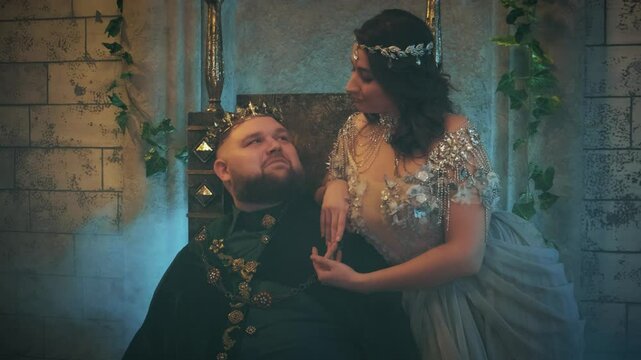 Fantasy Evil tyrannical man king sits medieval royal throne. Woman Queen, blue dress silver crown diadem. lady sitting near stroking man with hand. Fairy elf Girl dangerous princess old style castle