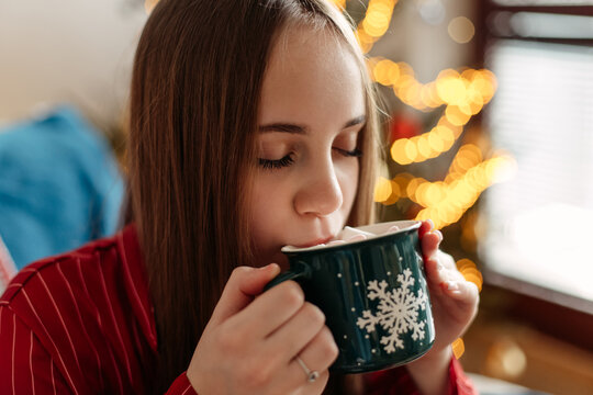 girl drinks cocoa with marshmallows in a green cup with snowflakes against the background of a Christmas tree