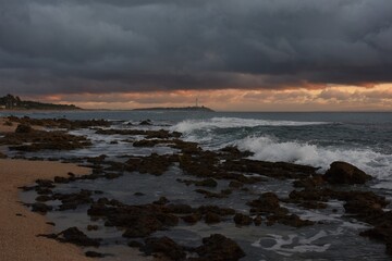 Obraz premium Wild waves rolling in at Playa de Zahora beach, Spain, massive dark threatening cloud bank in the back narrowing last light of the day to a stark pink edged strip around Faro de Trafalgar lighthouse