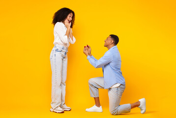 Loving black man on one knee holding open ring box, proposing to surprised lady against yellow background, side view, full length