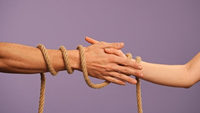 Man and woman hands tied together, tangled with rope, abusive or codependent relationships concept, isolated on purple studio background, close up shot.