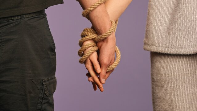 Man and woman holding hands tied together, tangled with rope, abusive unhealthy codependent relationships concept, isolated on purple studio background, close up shot.
