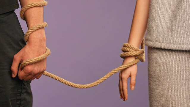 Man and woman hands tied together, tangled with rope, abusive unhealthy codependent relationships concept, isolated on purple studio background, close up shot.