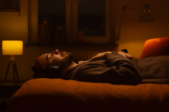A man lying on a bed wearing wireless headphones listening. - Powered by Adobe