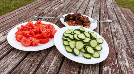 A picnic table with three white plates containing grilled meat, tomatoes and cucumbers.