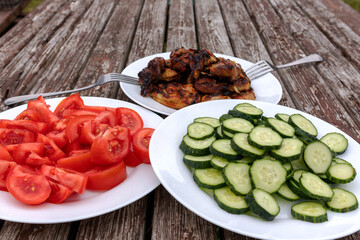 A picnic table with three white plates containing grilled meat, tomatoes and cucumbers.