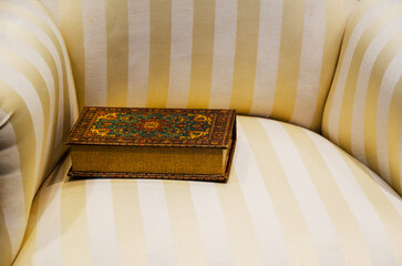 Old book in colorful leather binding lying on soft classic sofa with white and yellow stripes