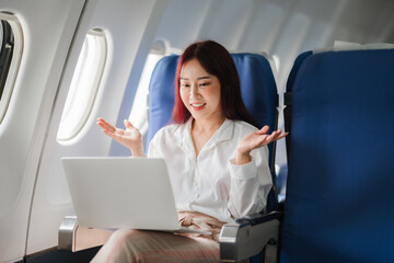 Business Woman on Airplane: A young, energetic woman in a white shirt smiles cheerfully as she uses...