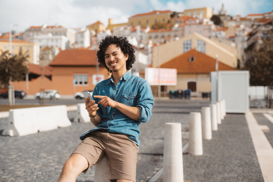 A cheerful biracial young man with curly hair sits casually on a white bollard outdoors in a sunny urban setting, holding a smartphone and smiling. Copy space area on the right