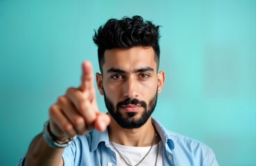 Obraz premium Confident young Lebanese man stares directly at camera. Points at eyes with determined look. Indoor studio shot against blue background. Man appears alert, focused, maybe spy someone watches for