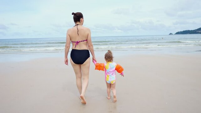 Mother leads little girl in inflatable armbands to foaming water edge
