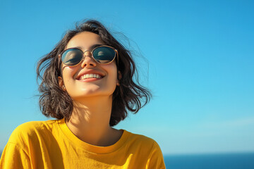 portrait of a happy young woman wearing sunglasses, with a blue sky and sea in the background. She is wearing a yellow shirt. This is a close-up portrait, taken on a sunny day with sunlight and sun ra