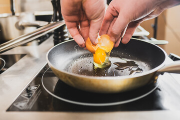 A close-up of hands cracking an egg into a frying pan on a modern stove. The shiny surface reflects the kitchen setting, showcasing a cooking process in action.