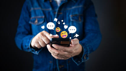 A person in a denim jacket interacts with a smartphone, surrounded by floating emojis, chat bubbles, hearts, and icons representing social engagement and network connectivity in the digital world.