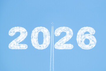 A plane flies high in a clear blue sky, trailing white smoke as it passes between clouds shaped like "2026." Copy space for New Year’s messages or creative designs