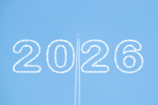A plane flies high in a clear blue sky, trailing white smoke as it passes between clouds shaped like "2026." Copy space for New Year&rsquo;s messages or creative designs