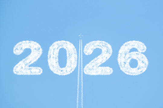 A plane flies high in a clear blue sky, trailing white smoke as it passes between clouds shaped like "2026." Copy space for New Year&rsquo;s messages or creative designs