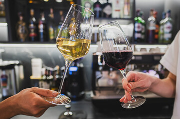 two glasseTwo hands clink glasses of white and red wine in a modern bar setting, with a blurred background of bottles. Cheers to celebration and moments shared.s of wine