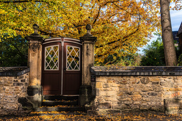 A sandstone wall with an ornate wooden door leads to a public garden with deciduous trees in warm autumn colours