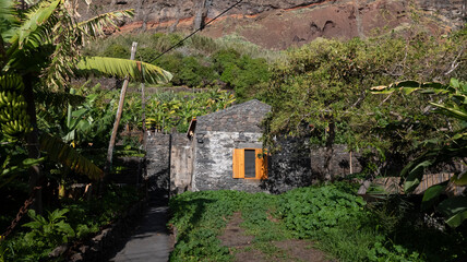 A small rustic stone house with bright orange shutters, surrounded by lush banana plants and greenery. Set against a rocky hillside, the scene exudes rural charm and tranquility.