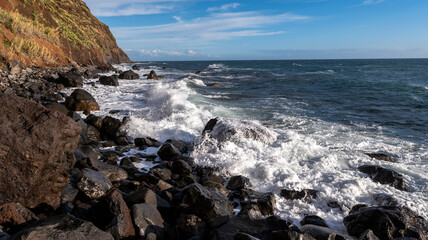 Waves crash against black volcanic rocks along a rugged coastline with steep cliffs covered in greenery. The bright blue sky and dynamic ocean create a dramatic and vibrant scene.