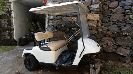 A white golf cart with beige seats parked near a rustic stone wall and a small garage. The scene blends modern utility with a rural, outdoor setting.