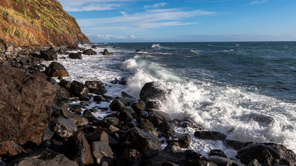 Waves crash against black volcanic rocks along a rugged coastline, with steep cliffs covered in greenery rising above. The bright blue sky highlights the dramatic coastal scenery.