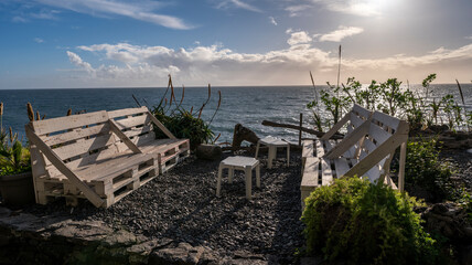 A cozy outdoor seating area made of white wooden pallet benches and plastic stools, overlooking the ocean. The setting features lush greenery and bright sunlight, creating a relaxing coastal ambiance.