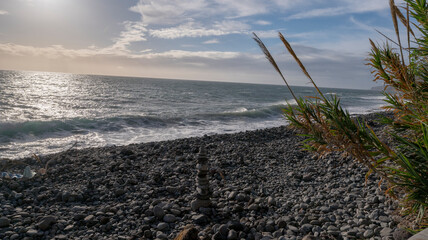 A rocky beach with gentle ocean waves glistening under the afternoon sun. The peaceful coastal setting features a clear sky and a stack of balanced stones in the foreground.