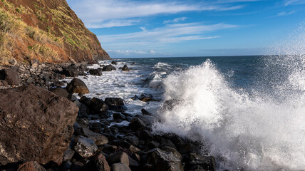 Waves crash against black volcanic rocks along a rugged coastline with steep cliffs covered in greenery. The vibrant ocean scene is illuminated by a bright blue sky.