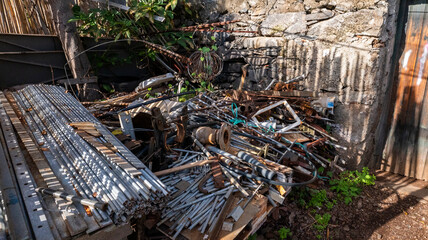 A pile of discarded metal scraps, pipes, and construction materials in a corner of an outdoor yard surrounded by a stone wall. The scene reflects industrial waste and recycling potential.