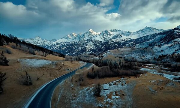 Scenic Mountain Landscape with Snow-Capped Peaks, Winding Road, and Open Fields Under a Dramatic Sky in a Serene Winter Setting