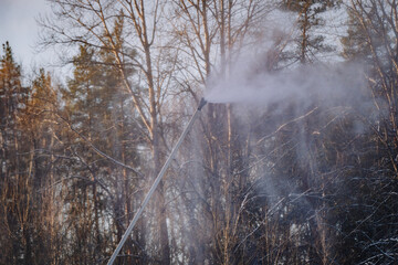 A snow cannon pipe sprays artificial snow into the air with a forest in the background during a cold winter day.