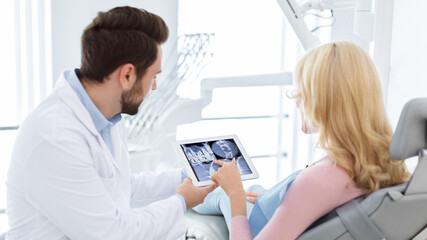 Female patient asking male dentist something, poiting at pad showing her jaws x-ray, modern dental clinic interior. Dentist and woman in dental chair discussing treatment, using digital tablet © Prostock-studio