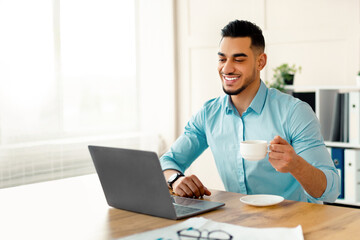 Fototapeta premium Happy young Arab man drinking coffee while using laptop at modern office. Handsome Eastern entrepreneur enjoying hot drink during online meeting with partner or colleague