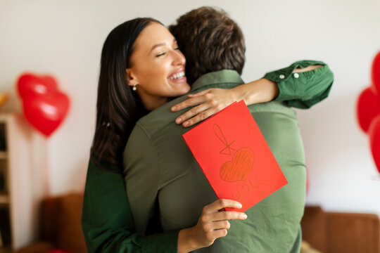 Valentine's Day. Happy woman holding greeting card and embracing beloved man, standing in living room decorated with balloons