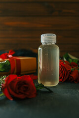Transparent bottle with clear liquid on a dark surface, surrounded by red roses and a gift box with a red ribbon, set against a wooden background for a warm, natural aesthetic