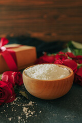 Wooden bowl filled with white bath salt, surrounded by vibrant red roses and scattered petals on a dark textured surface, creating a romantic and natural spa-like ambiance