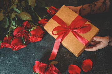 Close-up of hands holding a brown gift box tied with a red ribbon, surrounded by red roses and scattered petals on a dark textured surface. Romantic and elegant composition