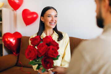 Young happy woman receiving beautiful bouquet of red roses from her boyfriend indoors, celebrating...