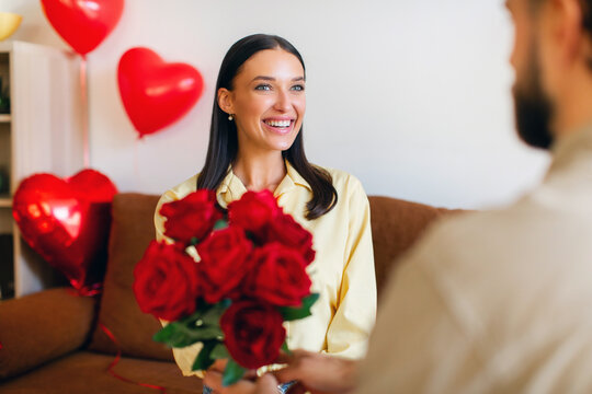Young happy woman receiving beautiful bouquet of red roses from her boyfriend indoors, celebrating Valentine's day together