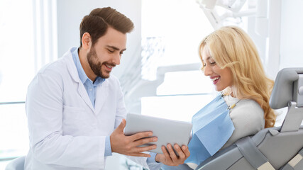 Smiling man dentist showing female patient jaw x-ray on digital tablet, dental clinic interior. Bearded man stomatologist having conversation with lady in dental chair, holding pad, side view